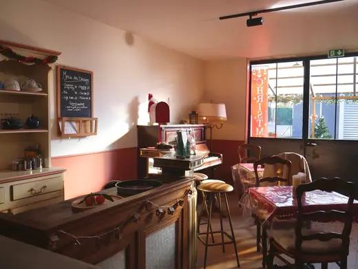 Le coin café : comptoir en bois, piano droit, tables nappées et menu des boissons à l'ardoise.