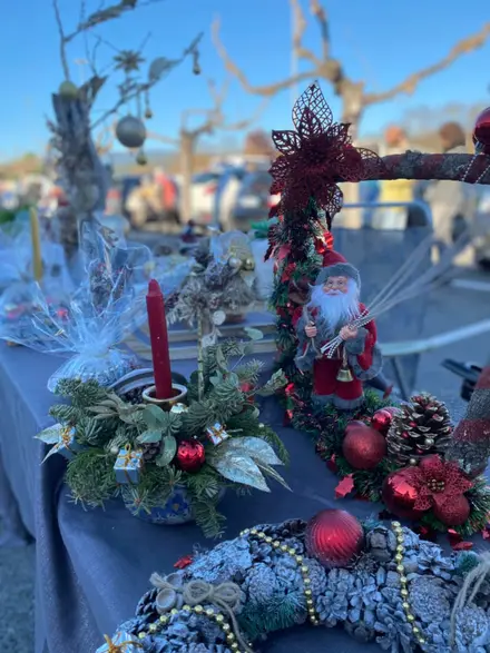 Un stand du marché de Noël : couronnes de pommes de pin, bougies et père Noël en tissu sous un ciel bleu.