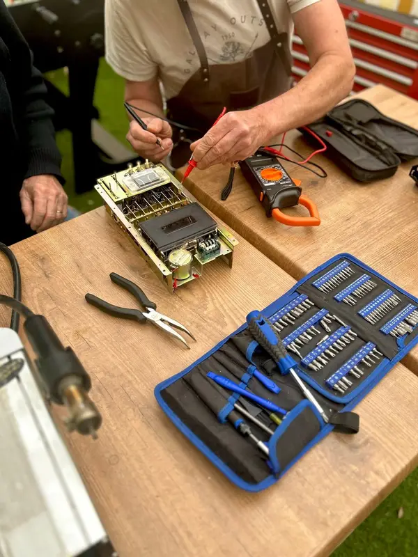 Un bénévole teste au multimètre l'alimentation d'un appareil au Repair Café, sous les yeux de sa propriétaire.