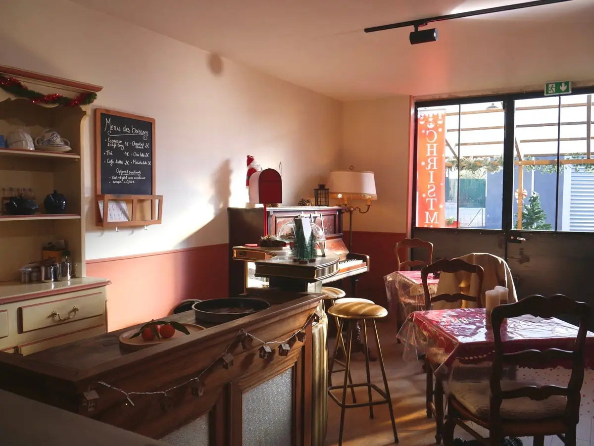 Le coin café : comptoir en bois, piano droit, tables nappées et menu des boissons à l'ardoise.
