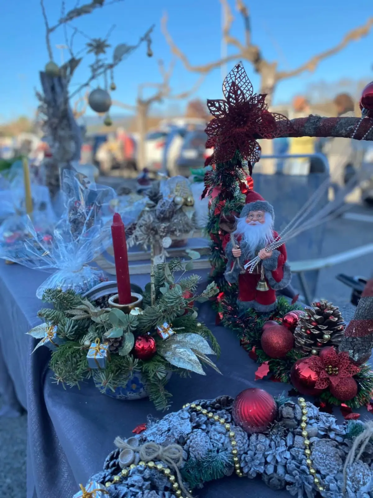 Un stand du marché de Noël : couronnes de pommes de pin, bougies et père Noël en tissu sous un ciel bleu.
